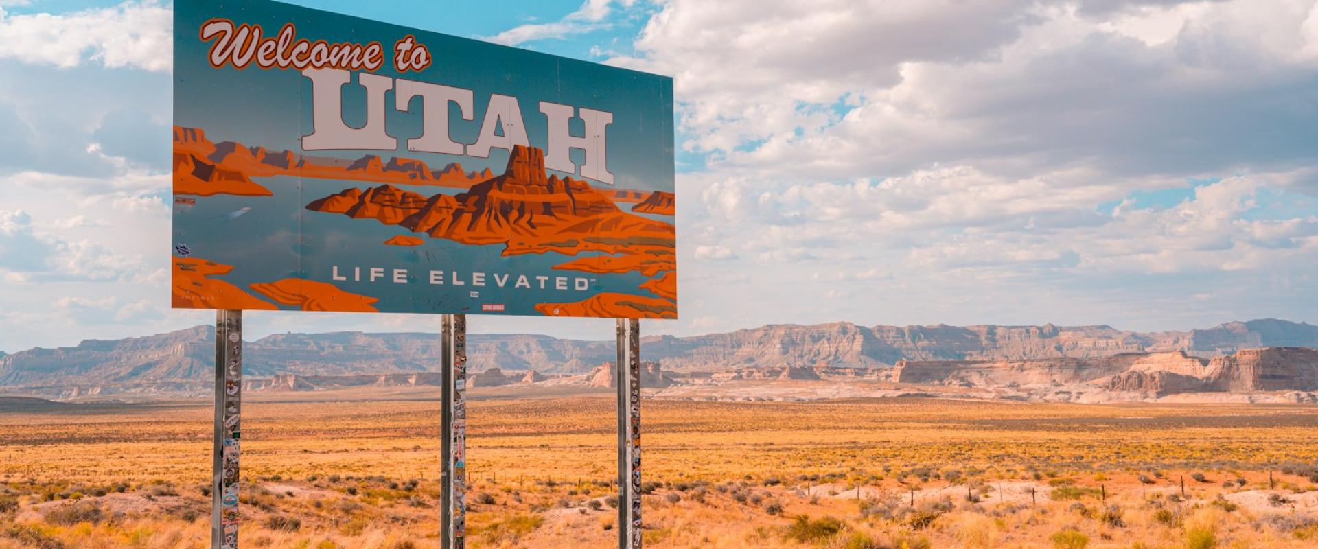 A large roadside billboard reading "Welcome to UTAH" with the slogan "Life Elevated" and an image of red rock formations, set against a desert landscape with mesas in the background.