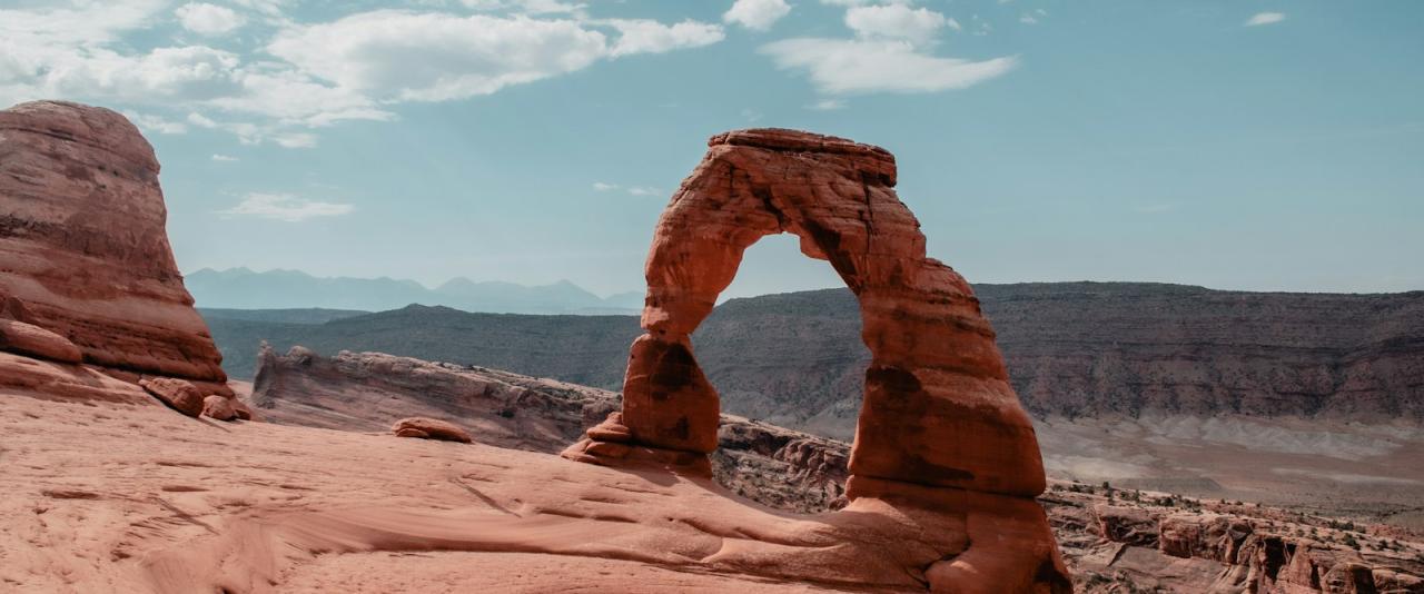 A large, freestanding natural sandstone arch situated on a rocky plateau with a desert canyon landscape and blue sky in the background.