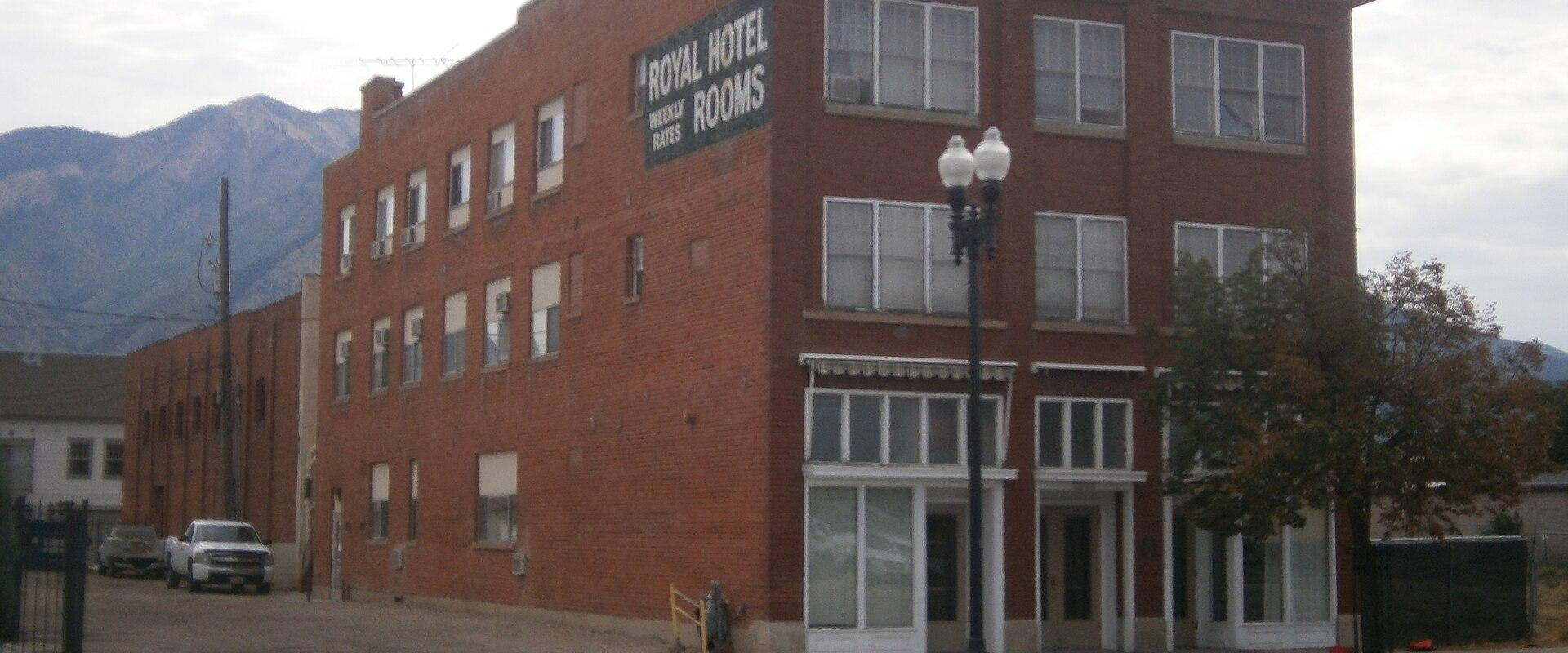 A multi-story red brick building with a painted "Royal Hotel" sign on its side, situated on a street corner with a mountain range visible in the background.