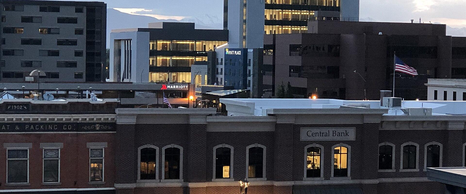 A view of a downtown streetscape at dusk, featuring a brick building with a "Central Bank" sign, a Marriott hotel, and a tall white office building, with the Wasatch Mountains visible in the background.