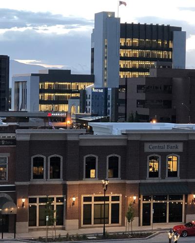 A view of a downtown streetscape at dusk, featuring a brick building with a "Central Bank" sign, a Marriott hotel, and a tall white office building, with the Wasatch Mountains visible in the background.