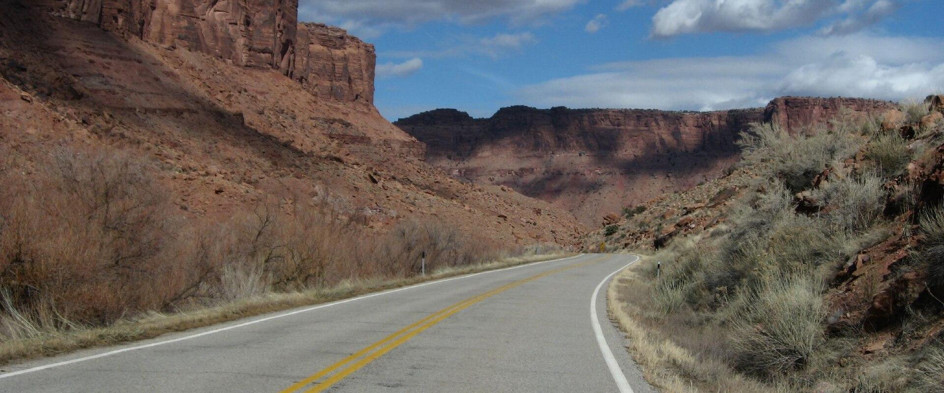A view from a vehicle looking out at a paved two-lane road curving to the right, flanked by red rock cliffs and desert scrub vegetation under a blue sky with clouds.