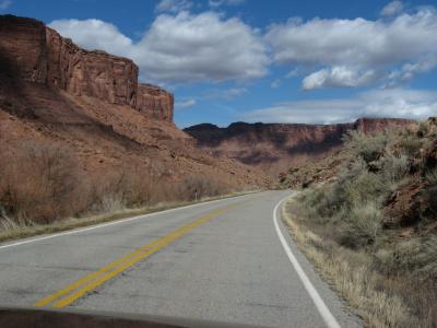 A view from a vehicle looking out at a paved two-lane road curving to the right, flanked by red rock cliffs and desert scrub vegetation under a blue sky with clouds.