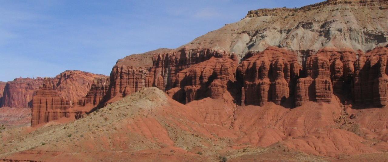 A wide landscape shot of red rock cliffs and a paved road with a silver SUV parked on the right side under a blue sky.