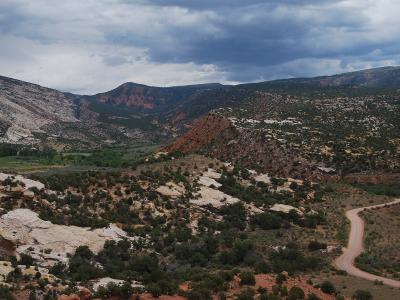 A wide landscape view showing a winding dirt road through a canyon with red rock formations, green vegetation, and a cloudy sky.