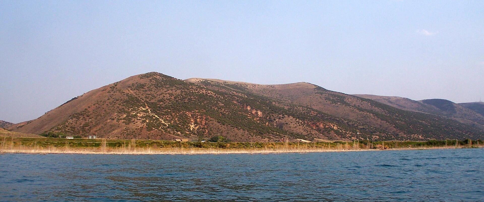A wide shot of a large body of water in the foreground, with a long, low hill or ridge covered in sparse, brownish-green vegetation along the far shore, set against a clear blue sky.