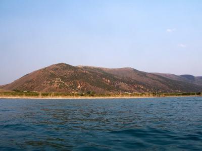 A wide shot of a large body of water in the foreground, with a long, low hill or ridge covered in sparse, brownish-green vegetation along the far shore, set against a clear blue sky.