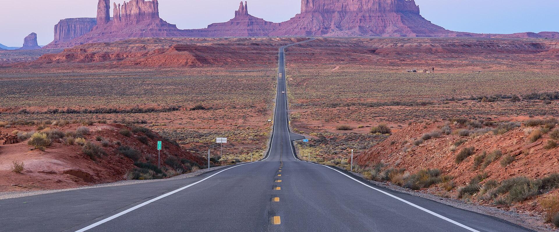 A paved two-lane road stretching straight into the distance, flanked by desert scrub, with the iconic red rock buttes of the Monument Valley Navajo Tribal Park visible in the background.