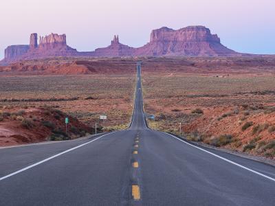 A paved two-lane road stretching straight into the distance, flanked by desert scrub, with the iconic red rock buttes of the Monument Valley Navajo Tribal Park visible in the background.