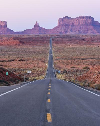 A paved two-lane road stretching straight into the distance, flanked by desert scrub, with the iconic red rock buttes of the Monument Valley Navajo Tribal Park visible in the background.