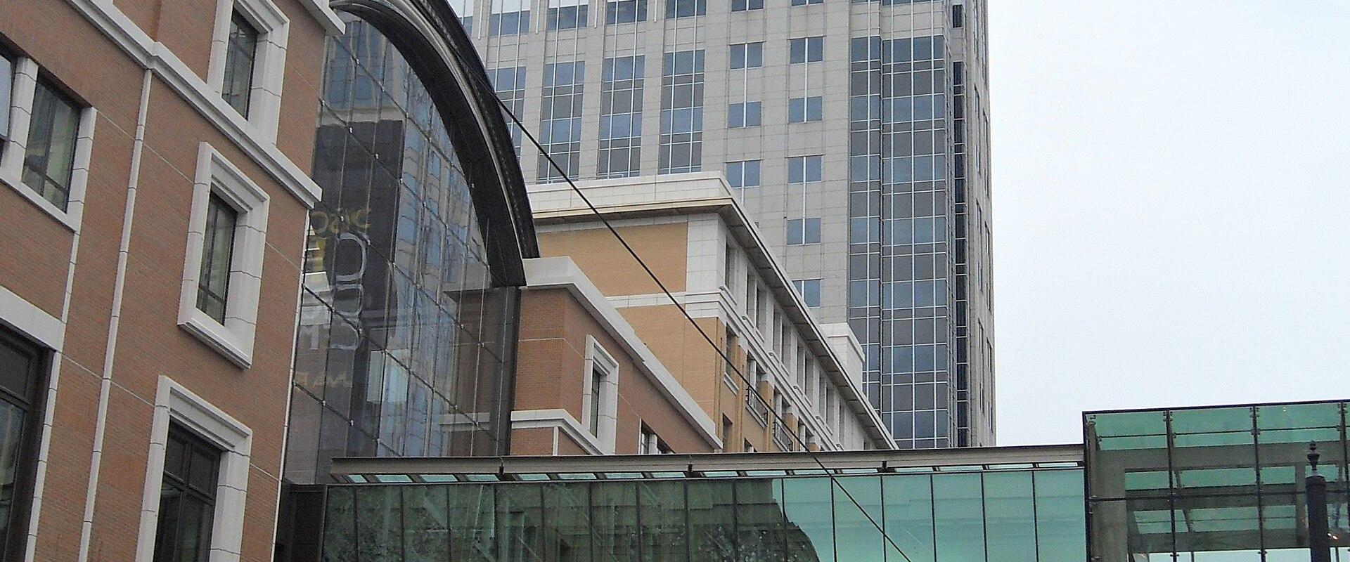 A street-level view of an urban pedestrian bridge with glass railings and the word "CITY" visible on its side, set against brick buildings and a taller office tower in the background.