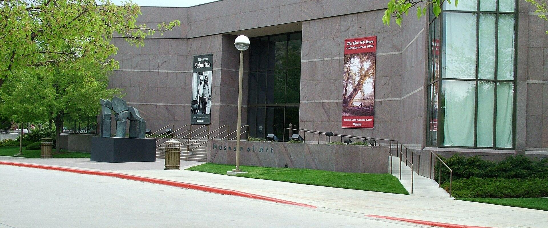 The image shows the exterior of a modern building with a low, horizontal profile, featuring a large glass facade on the right and a solid, light-colored stone wall on the left. A prominent sculpture of a dark, abstract figure stands on a black pedest