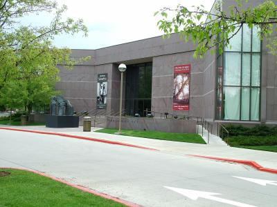 The image shows the exterior of a modern building with a low, horizontal profile, featuring a large glass facade on the right and a solid, light-colored stone wall on the left. A prominent sculpture of a dark, abstract figure stands on a black pedest