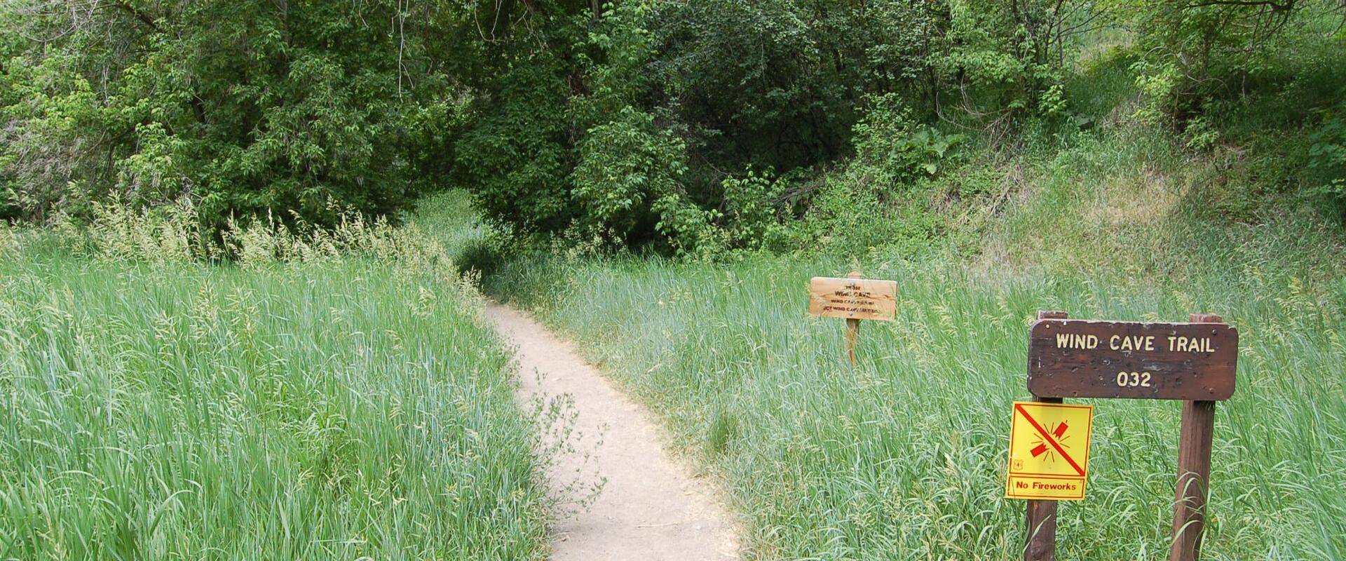 A dirt trail winding through tall green grass and trees, with a wooden sign on the right that reads "WIND CAVE TRAIL 032" and a yellow "No Fireworks" sign attached to it.