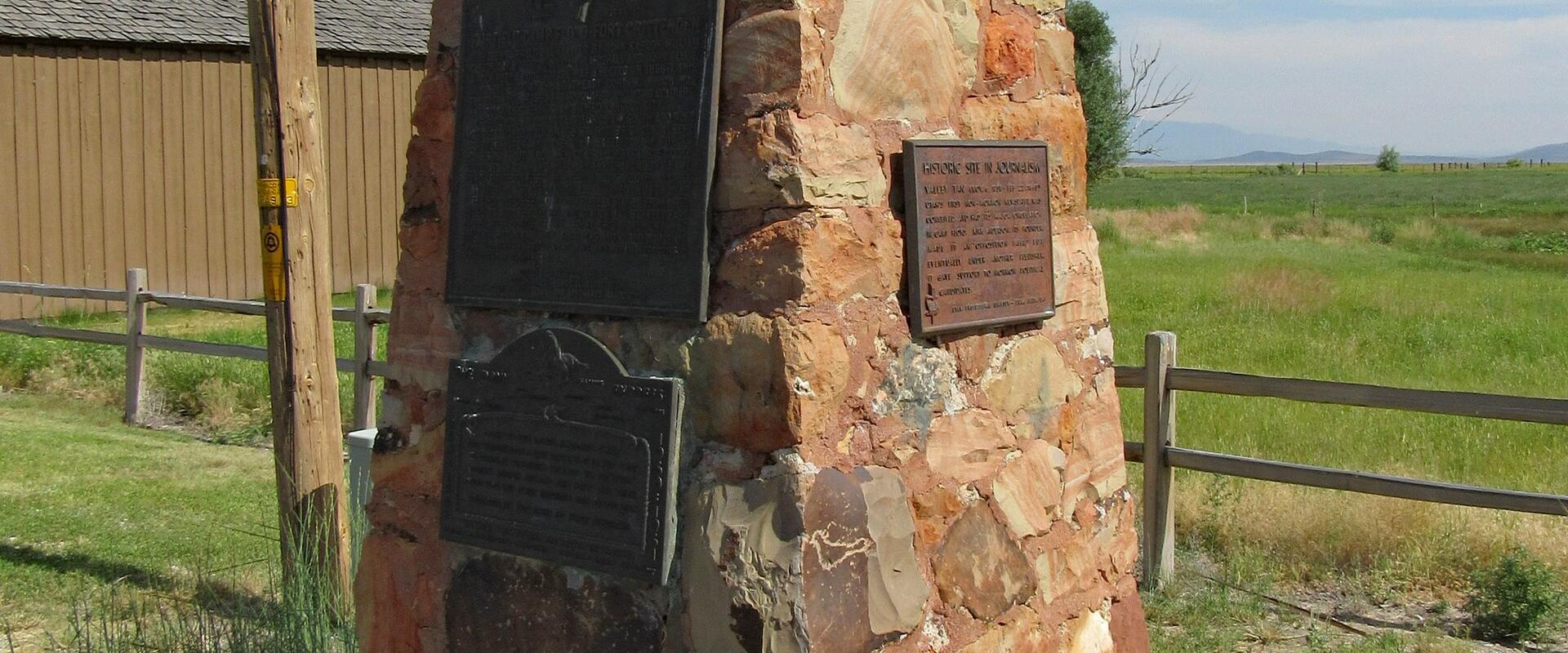 A stone obelisk monument featuring several bronze plaques, including one clearly reading "CAMP FLOYD 1858-1860", situated next to a wooden fence and a building in a rural setting.
