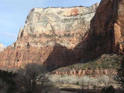 A landscape photograph showing a large, stratified sandstone cliff face (likely the Navajo Sandstone) with a shadow cast across its face, above a foreground of bare, wintry trees and a dry wash.