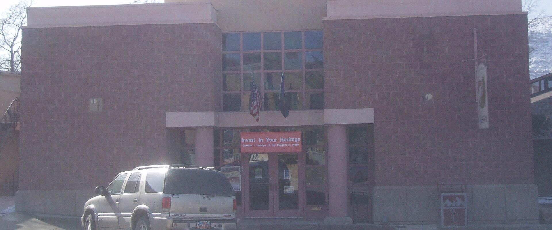 The image shows the exterior facade of a single-story building with a beige stucco upper section and a reddish-brown brick lower section, featuring a central entrance with glass doors, a large window, and the text "DAN O'L AURIE MUSEUM" mounted above
