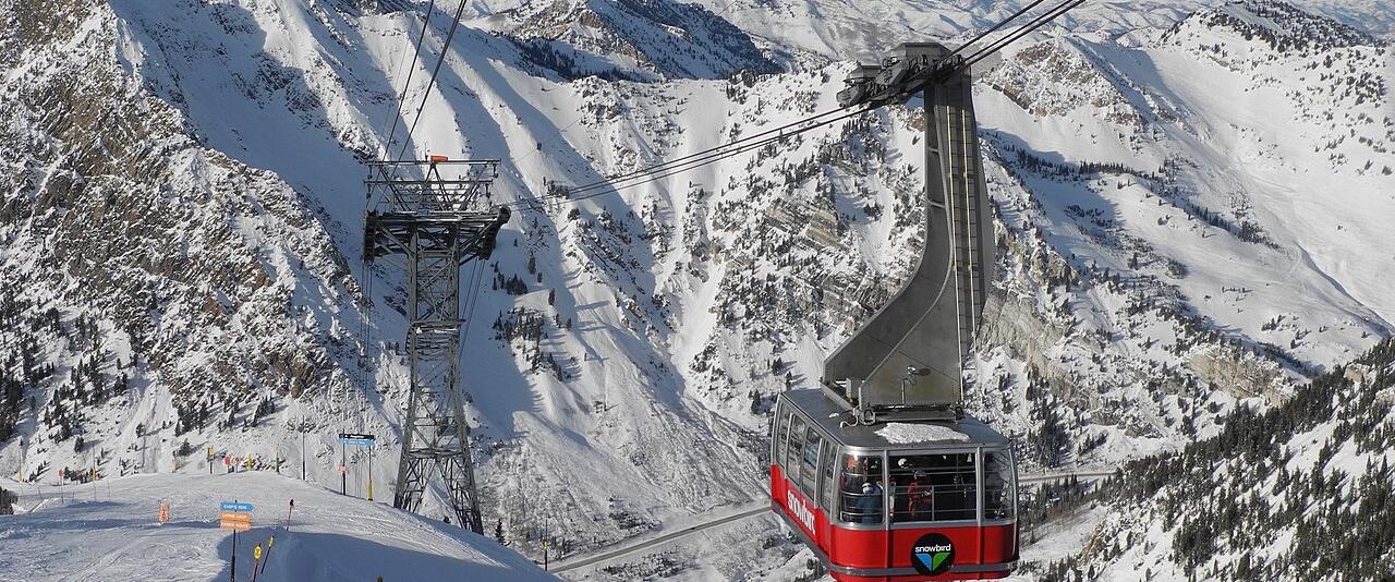 The red Snowbird Aerial Tram cabin above snowy slopes, with the Wasatch peaks of Little Cottonwood Canyon behind