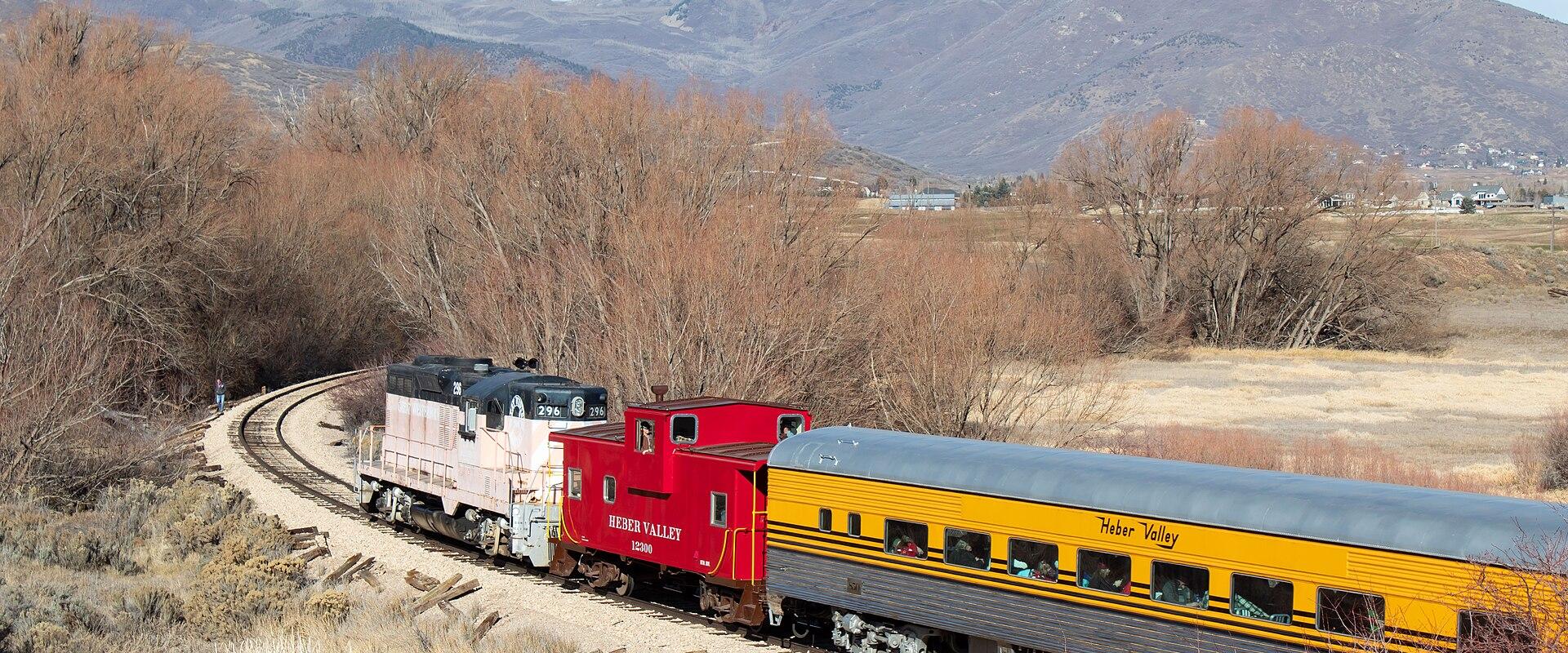 The image shows a train with a white and black locomotive, a red caboose, and a yellow passenger car traveling along a track through a dry, grassy landscape with mountains in the background.