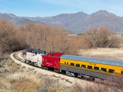 The image shows a train with a white and black locomotive, a red caboose, and a yellow passenger car traveling along a track through a dry, grassy landscape with mountains in the background.