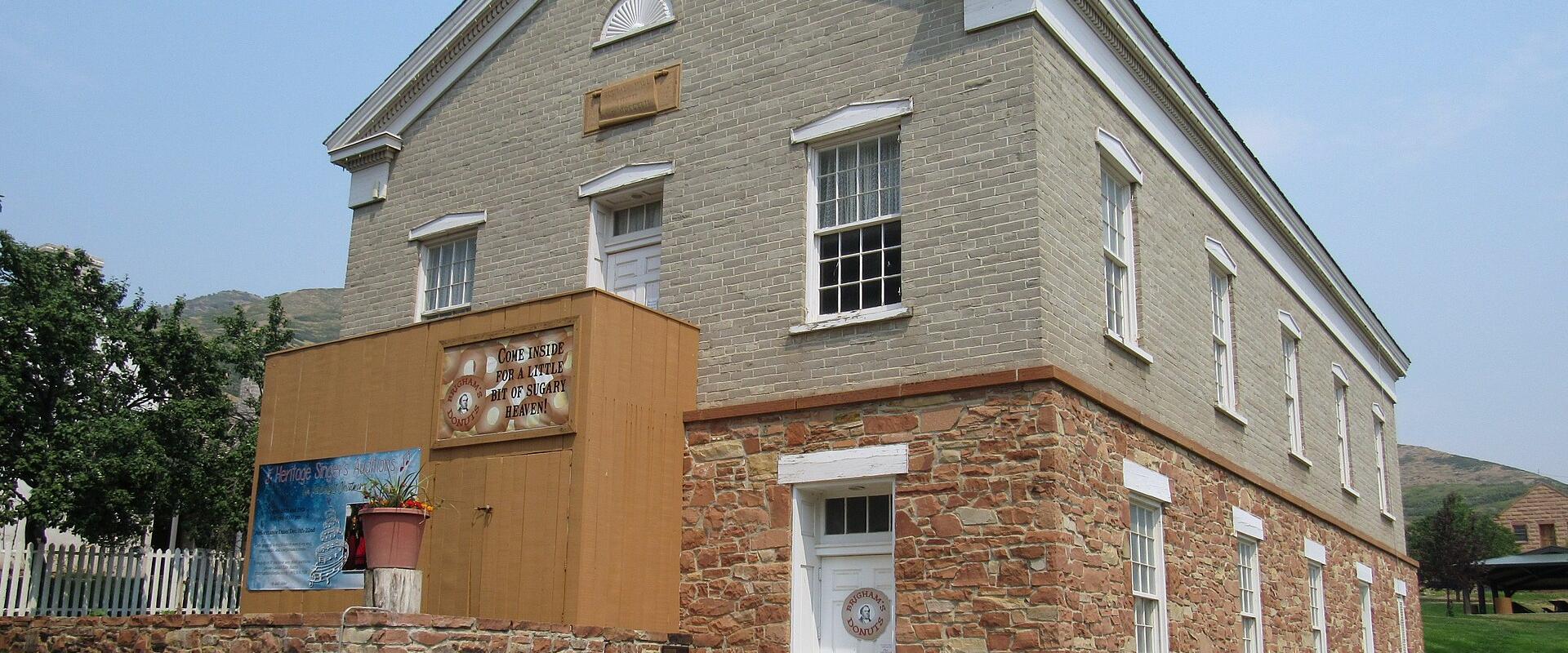 A two-story building with a light-colored brick facade and a stone foundation, featuring a temporary wooden structure attached to its side, set against a clear sky.