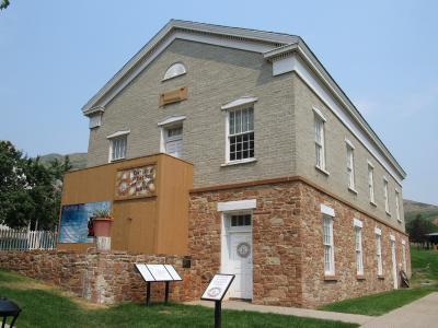 A two-story building with a light-colored brick facade and a stone foundation, featuring a temporary wooden structure attached to its side, set against a clear sky.