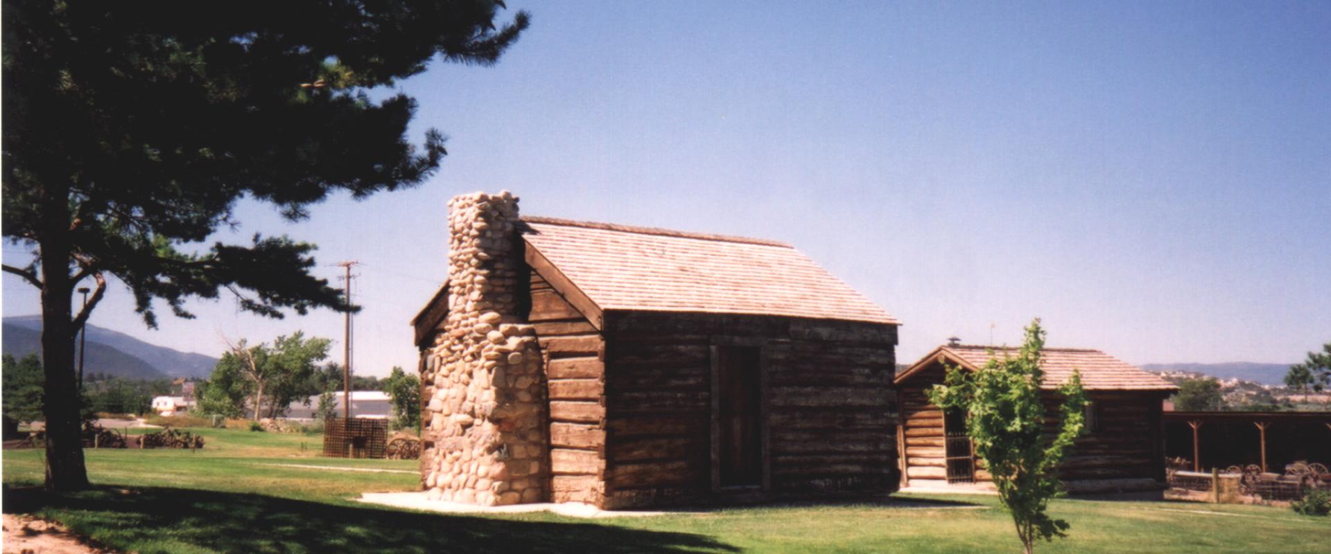 A log cabin with a stone chimney and a wooden shingle roof, situated on a grassy lawn with a large pine tree to the left and another building visible in the background.