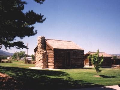 A log cabin with a stone chimney and a wooden shingle roof, situated on a grassy lawn with a large pine tree to the left and another building visible in the background.