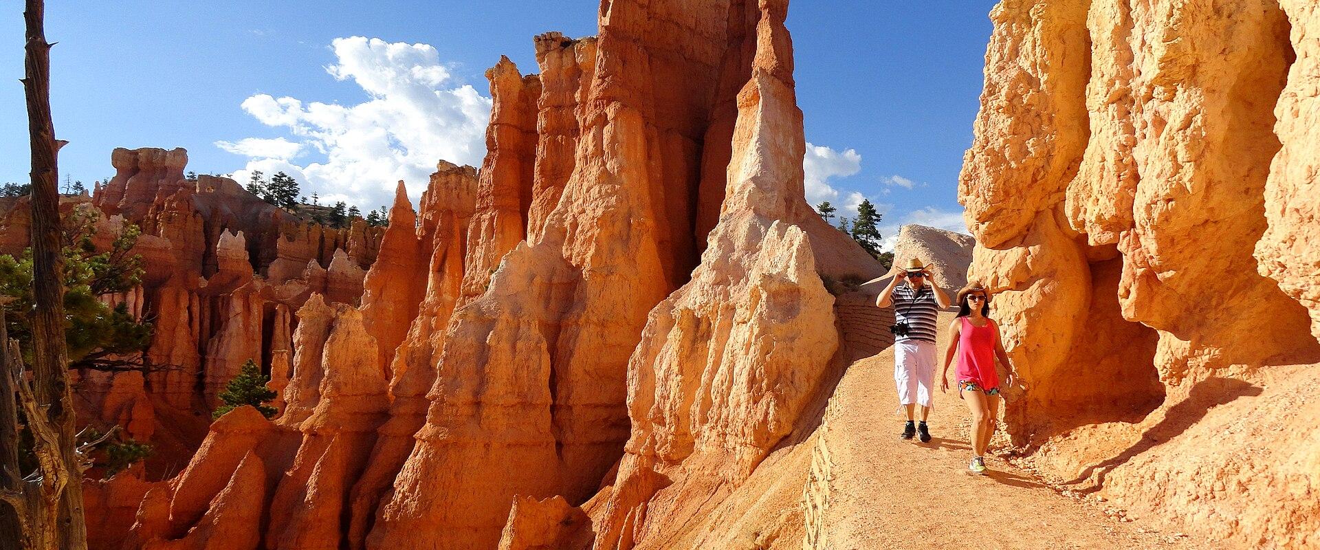 The image shows a dirt trail winding through a canyon of tall, orange-red rock spires (hoodoos) with two people walking along the path, set against a blue sky.