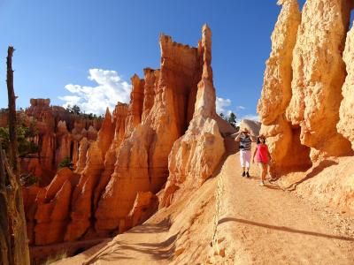 The image shows a dirt trail winding through a canyon of tall, orange-red rock spires (hoodoos) with two people walking along the path, set against a blue sky.