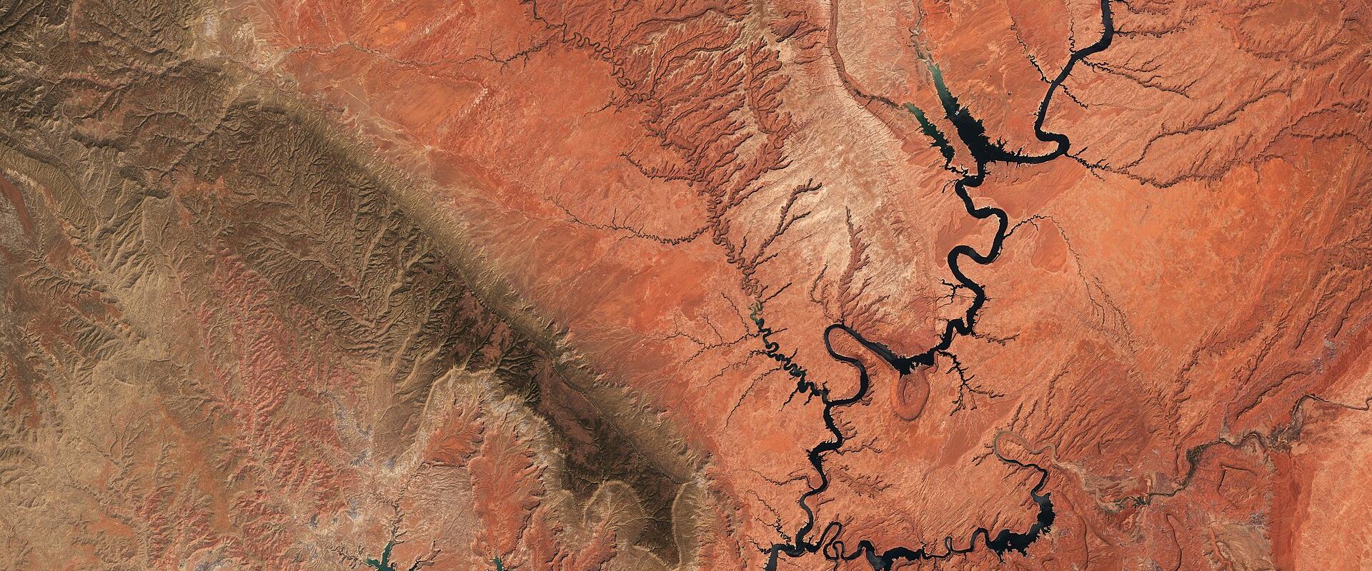 A satellite/aerial view of a winding, dark-water reservoir cutting through red desert terrain, with a prominent conical volcanic cinder cone visible in the lower center.