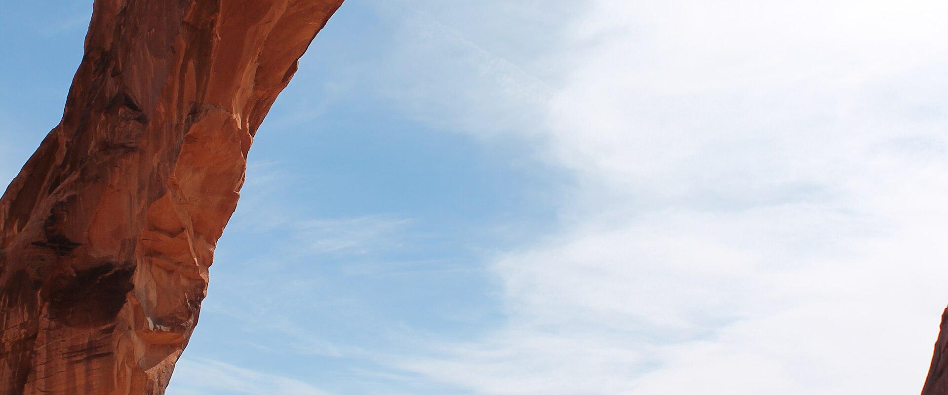 A large, reddish-brown natural sandstone arch with a wide span, set against a blue sky with light clouds, overlooking a desert landscape with distant mesas.