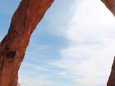 A large, reddish-brown natural sandstone arch with a wide span, set against a blue sky with light clouds, overlooking a desert landscape with distant mesas.