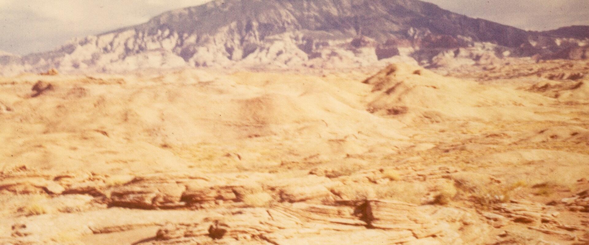 A vintage photograph showing a desert landscape with a large, flat-topped mesa (likely Navajo Mountain) in the background, a sandy foreground, and a small, dark, tent-like structure in the lower right.
