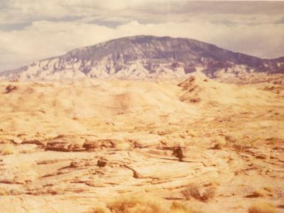 A vintage photograph showing a desert landscape with a large, flat-topped mesa (likely Navajo Mountain) in the background, a sandy foreground, and a small, dark, tent-like structure in the lower right.