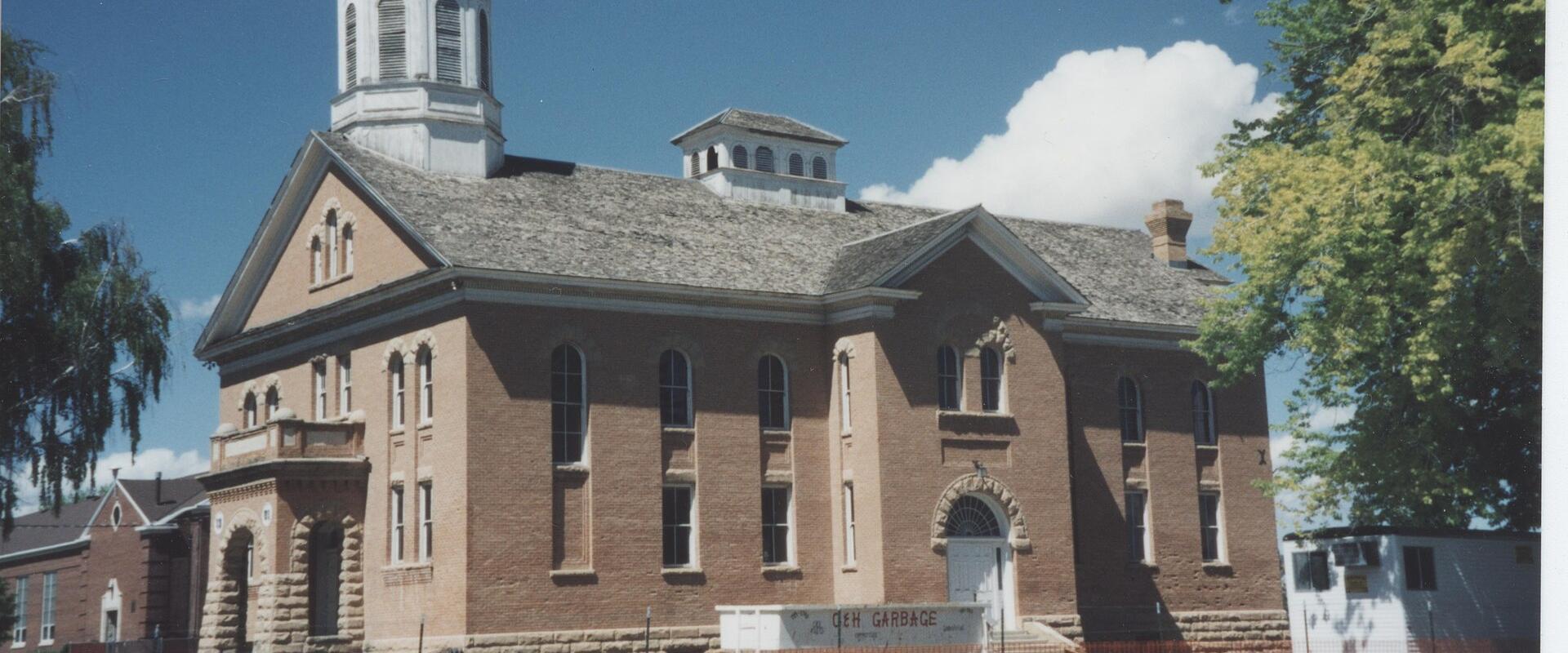A large, two-story brick building with a central white cupola and a smaller side cup, featuring a white front door and a dumpster in the foreground.