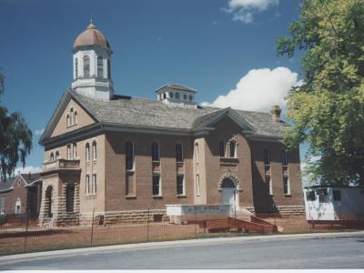 A large, two-story brick building with a central white cupola and a smaller side cup, featuring a white front door and a dumpster in the foreground.