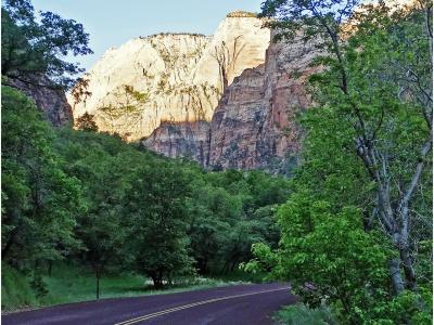 A paved two-lane road curves through a lush, green, tree-lined area, with a large, light-colored sandstone cliff face rising in the background.