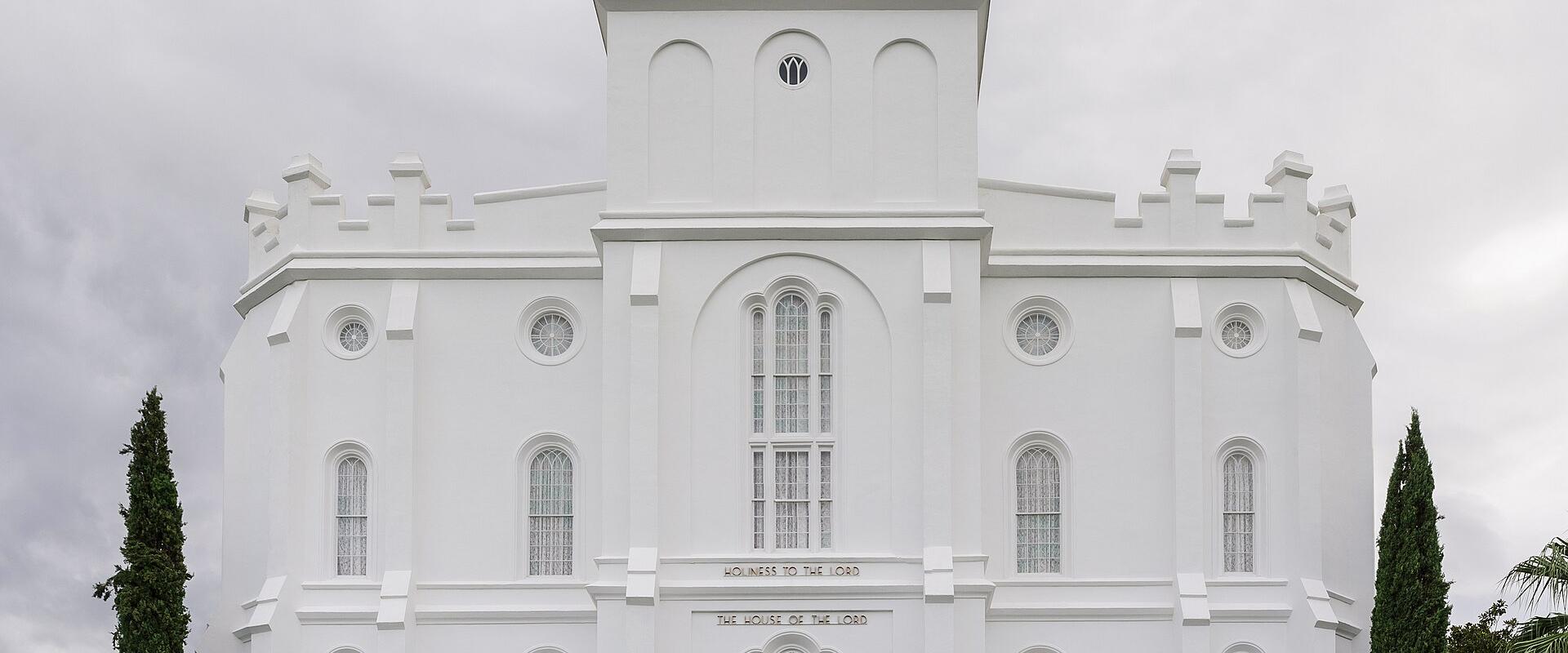 A white, multi-tiered temple building with a central spire, flanked by palm trees and manicured lawns, with people walking on the grounds.