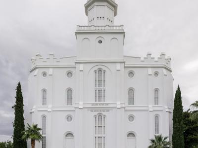 A white, multi-tiered temple building with a central spire, flanked by palm trees and manicured lawns, with people walking on the grounds.