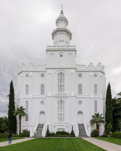 A white, multi-tiered temple building with a central spire, flanked by palm trees and manicured lawns, with people walking on the grounds.