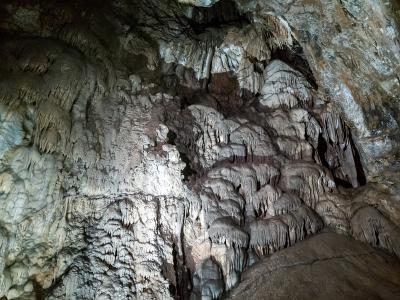 The image shows the interior of a limestone cave, featuring close-up views of stalactites and other mineral formations illuminated by artificial light.