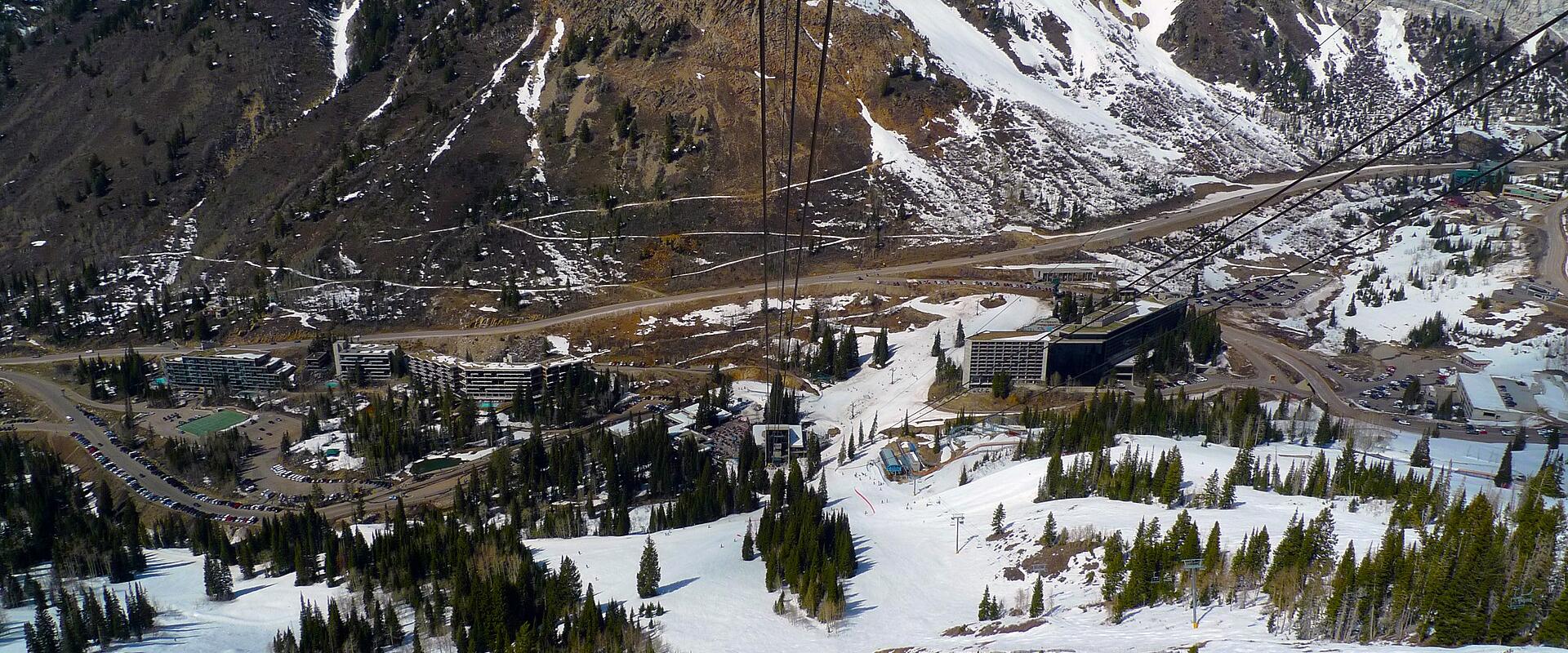 A high-angle view looking down from a ski lift onto a large, multi-story hotel complex (the Jughandle) and surrounding lodge buildings, with steep, snow-dusted mountain slopes in the background.