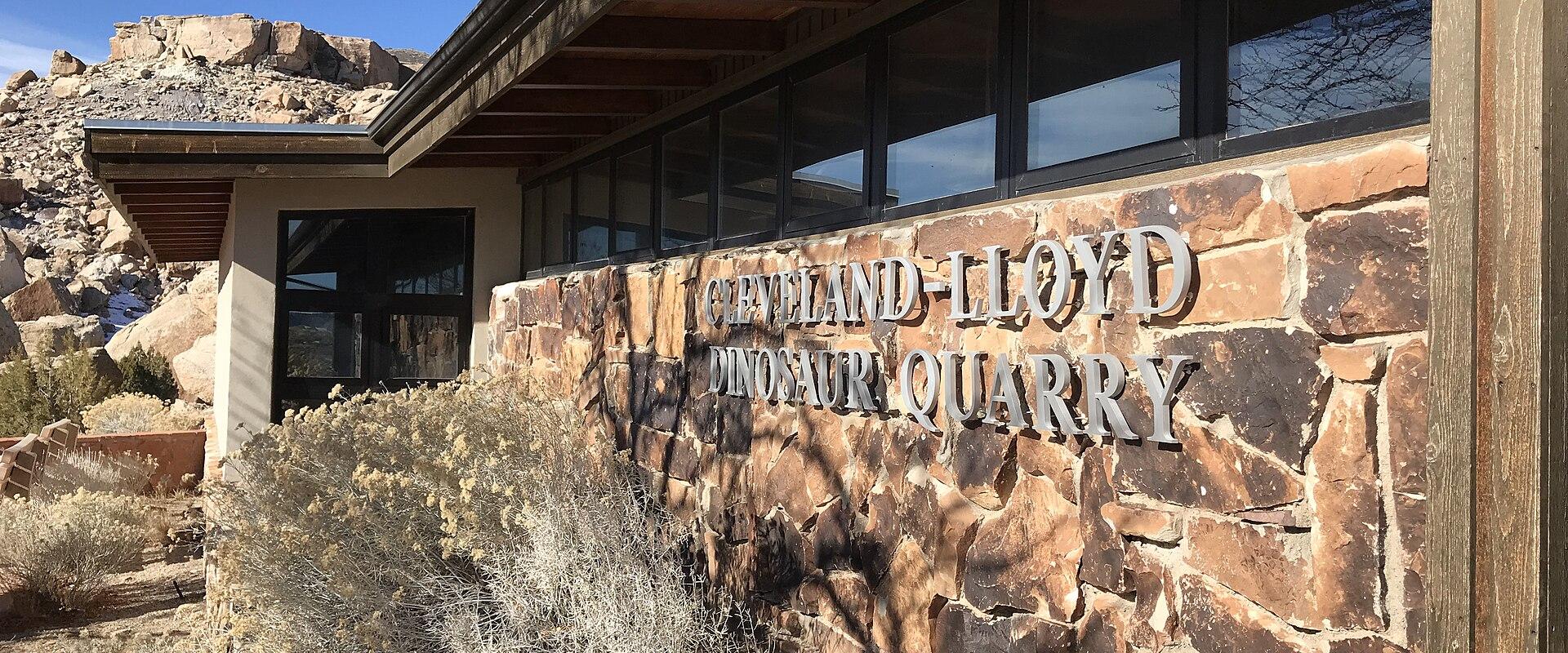 The image shows the exterior of a building with a stone facade, featuring large metal lettering that reads "CLEVELAND LLOYD DINOSAUR QUARRY" mounted on the wall, with desert vegetation in the foreground.