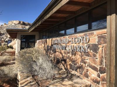 The image shows the exterior of a building with a stone facade, featuring large metal lettering that reads "CLEVELAND LLOYD DINOSAUR QUARRY" mounted on the wall, with desert vegetation in the foreground.