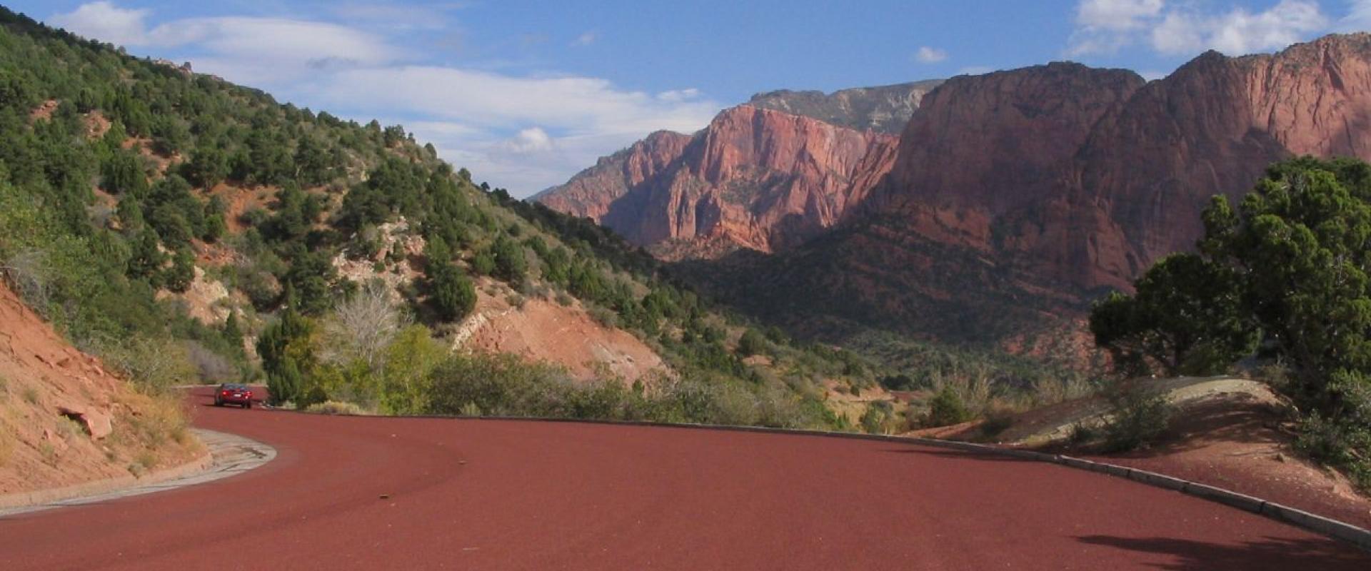 The image shows a paved road with a distinct reddish-brown surface winding through a landscape of steep, red rock cliffs and green vegetation, with a red car visible in the distance.