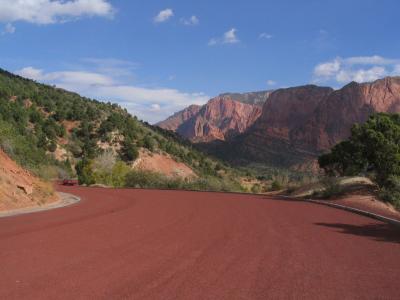 The image shows a paved road with a distinct reddish-brown surface winding through a landscape of steep, red rock cliffs and green vegetation, with a red car visible in the distance.