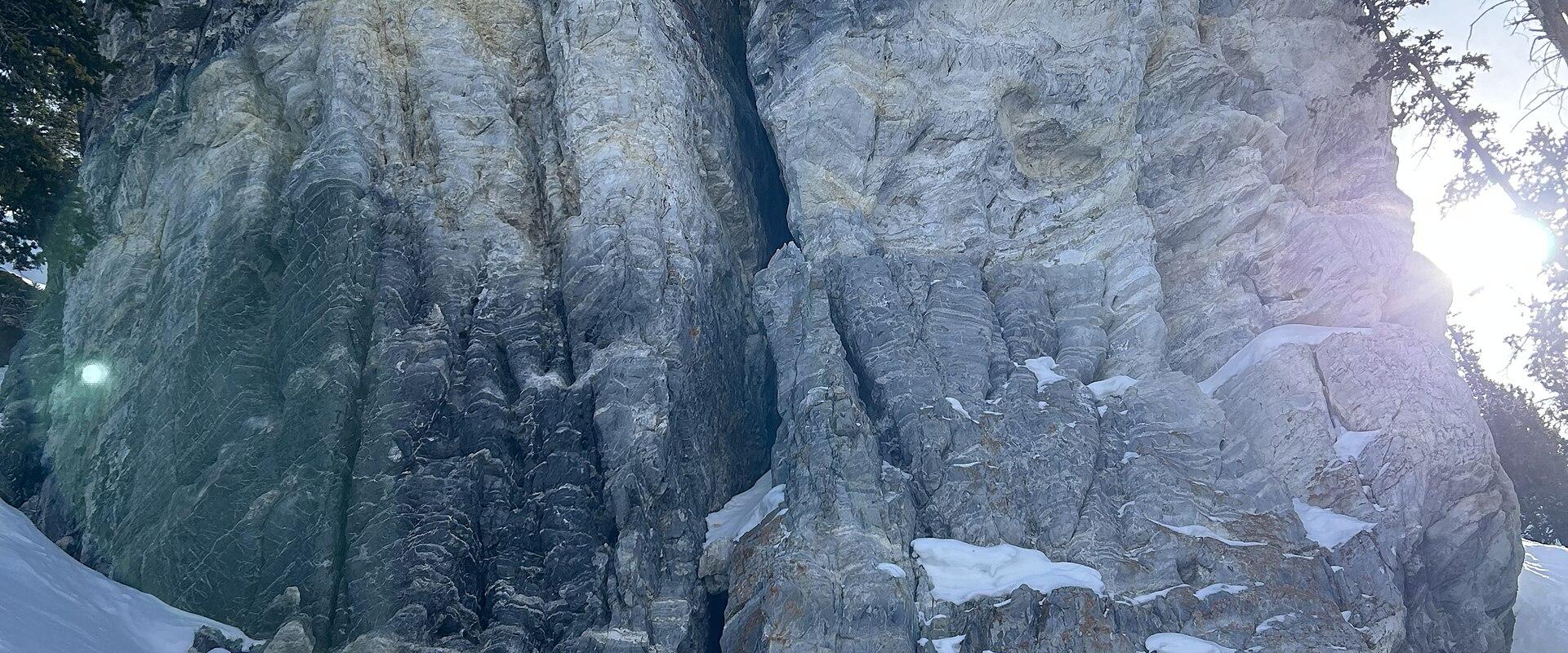 A low-angle, close-up view of a large, grey, craggy rock formation with a vertical fissure, set against a blue sky, with snow on the ground in the foreground and evergreen trees visible at the edges.