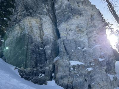 A low-angle, close-up view of a large, grey, craggy rock formation with a vertical fissure, set against a blue sky, with snow on the ground in the foreground and evergreen trees visible at the edges.