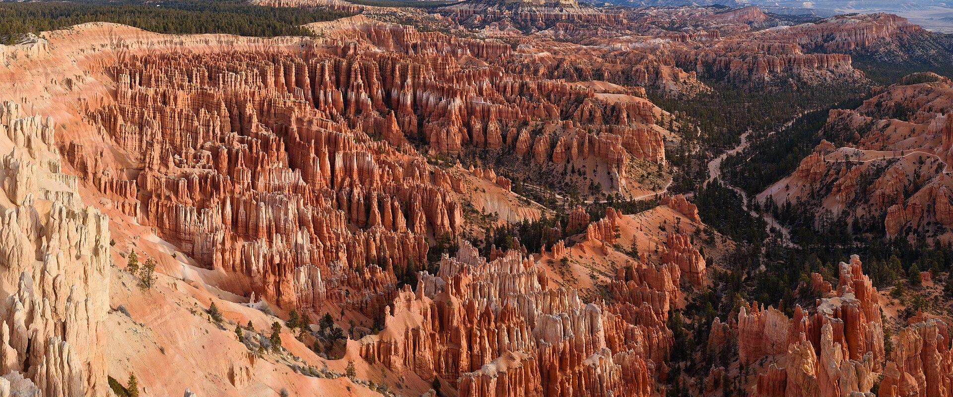 A panoramic view of a deep canyon amphitheater filled with numerous tall, thin, orange-red rock spires (hoodoos) and scattered green coniferous trees, set against a blue sky.